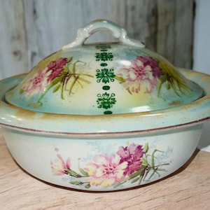 Puede incluir: Un mantequillero de cerámica vintage con tapa. La fuente está decorada con un patrón floral en tonos rosa, verde y amarillo. La tapa tiene un asa y también está decorada con flores.