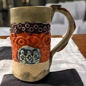 May include: A ceramic mug with a brown, orange, and white design. The mug features three monkeys with their hands covering their eyes, ears, and mouth. The mug has a textured surface and a handle.
