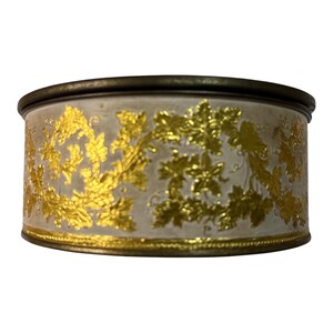 May include: A round, antique-style container with a cream-colored body and gold leaf grape vine design. The container has a dark bronze-colored rim and base. The gold leaf design wraps around the container.
