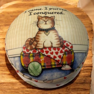 May include: A round decorative box with a cat theme. The lid features a tabby cat sitting on a bed with a red heart-patterned blanket. The text "I came. I purred. I conquered." is printed on the lid. A green yarn ball and a toy mouse are also visible.