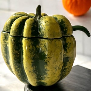 May include: A green and yellow ceramic pumpkin shaped sugar bowl with a lid and a small handle. The bowl is decorated with a mottled green and yellow glaze.