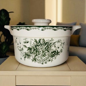 May include: A white ceramic casserole dish with a lid, featuring a green floral pattern. The dish has two handles and a decorative green border. The design is a classic, vintage style.