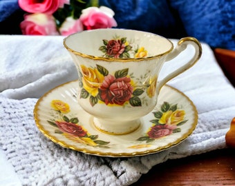 May include: Cream-colored teacup and saucer set with a floral rose design in yellow and red. The teacup and saucer have gold trim. The teacup is on a white surface with pink roses in the background.