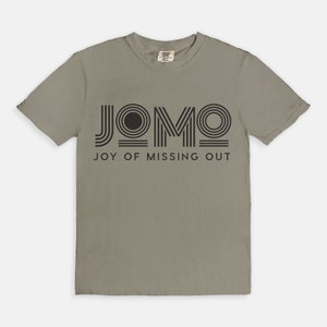 Puede incluir: Una camiseta gris claro con un estampado gráfico negro que dice "JOMO" en una fuente estilizada. El texto debajo del gráfico dice "JOY OF MISSING OUT".