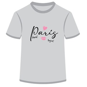 Puede incluir: Una camiseta gris con un gráfico negro que dice "París" con un corazón rosa y labios. El texto también dice "¡Amour!" y "¡Bonjour!"