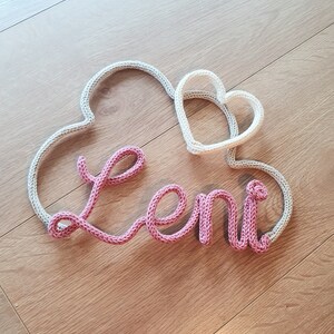 May include: A knitted name sign with the name "Leni" in pink yarn and two hearts in beige yarn.