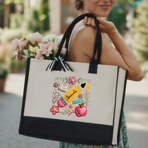 May include: Cream-colored tote bag with black trim and handles. The bag features an embroidered design of a pencil, apple, open book, and flowers. The name "Amanda" is written on the pencil. The bag is filled with pink and white flowers.