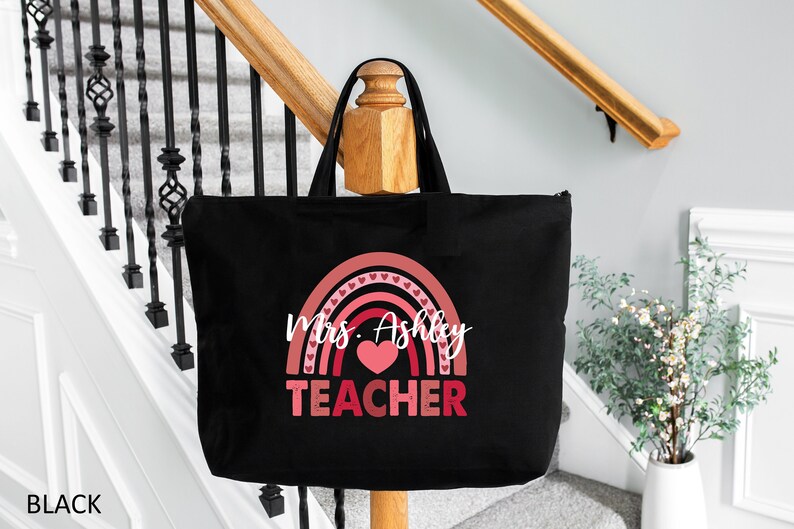K&ouml;nnte beinhalten: Schwarze Tragetasche mit einem rosafarbenen Regenbogen-Design und dem Text "Mrs. Ashley TEACHER" in Wei&szlig;.
