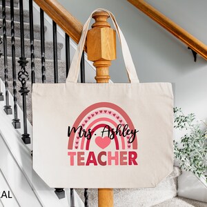 K&ouml;nnte beinhalten: Eine naturfarbene Canvas-Tragetasche mit einem rosafarbenen Regenbogen-Design und dem aufgedruckten Text "Mrs. Ashley TEACHER".