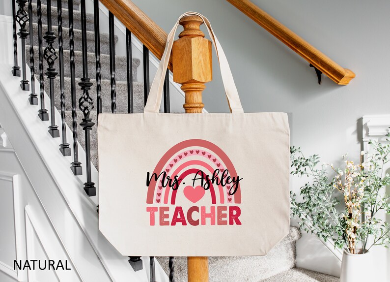 K&ouml;nnte beinhalten: Eine naturfarbene Canvas-Tragetasche mit einem rosafarbenen Regenbogen-Design und dem aufgedruckten Text "Mrs. Ashley TEACHER".
