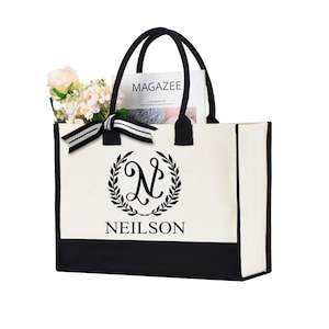 May include: A white canvas tote bag with black trim and a black and white monogram design. The monogram features a stylized letter "N" inside a wreath. The text "NEILSON" is printed below the monogram. The bag is shown with a magazine and flowers inside.