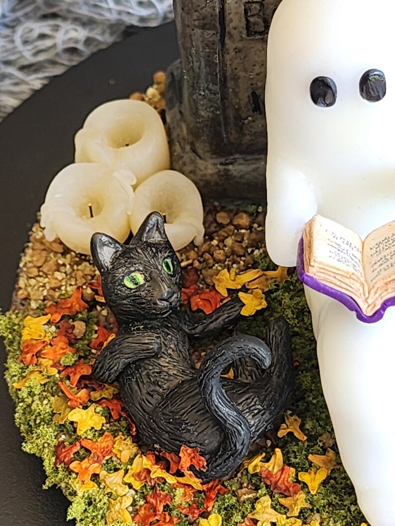 Close up of cat laying on back in autumn leaves. Ghost sitting beside gravestone reading a book with 2 black cats playing in the fall leaves. Halloween decor that will glow in the dark and has flickering candles.