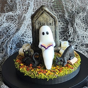 Ghost sitting beside gravestone reading a book with 2 black cats playing in the fall leaves. Halloween decor that will glow in the dark and has flickering candles.