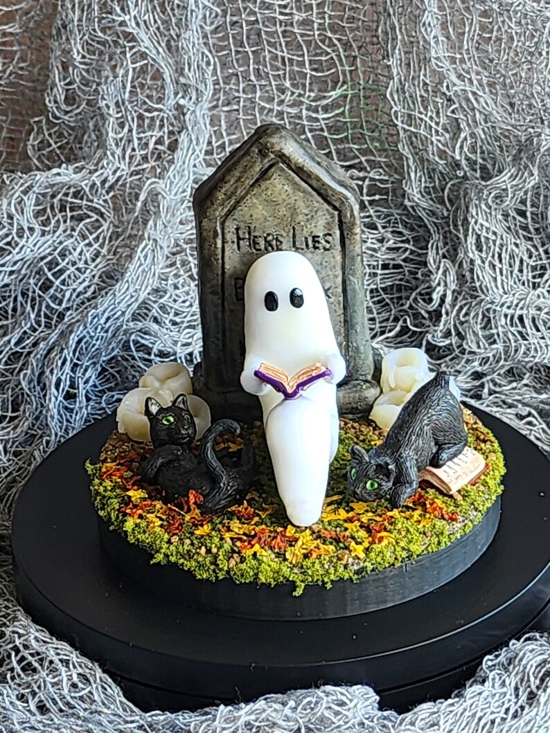 Ghost sitting beside gravestone reading a book with 2 black cats playing in the fall leaves. Halloween decor that will glow in the dark and has flickering candles.
