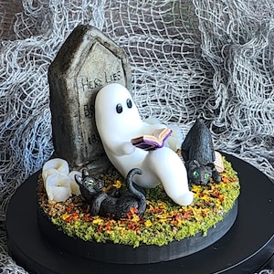 Side view to show detail. Ghost sitting beside gravestone reading a book with 2 black cats playing in the fall leaves. Halloween decor that will glow in the dark and has flickering candles.