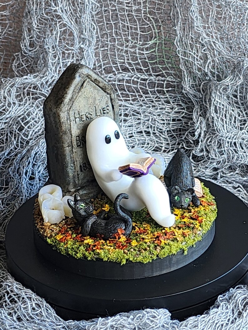 Side view to show detail. Ghost sitting beside gravestone reading a book with 2 black cats playing in the fall leaves. Halloween decor that will glow in the dark and has flickering candles.