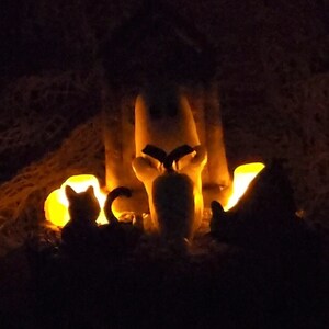 Dark photo to show flickering candles. Ghost sitting beside gravestone reading a book with 2 black cats playing in the fall leaves. Halloween decor that will glow in the dark and has flickering candles.