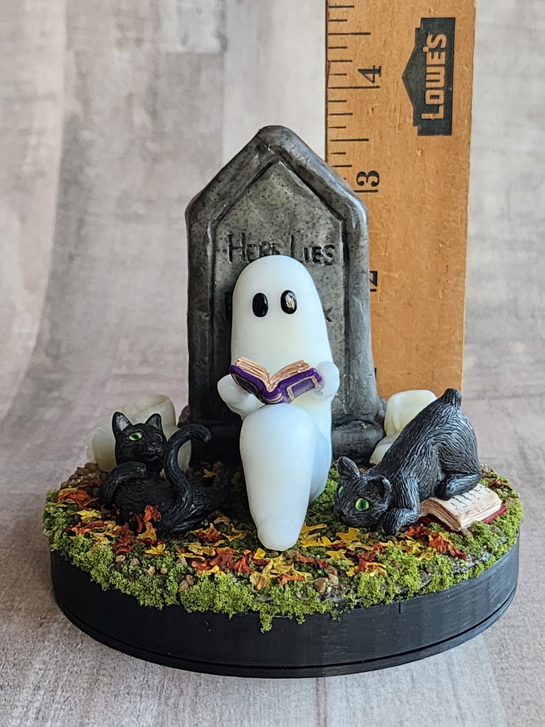 Sculpt is approximately 3.75 inches tall. Ghost sitting beside gravestone reading a book with 2 black cats playing in the fall leaves. Halloween decor that will glow in the dark and has flickering candles.