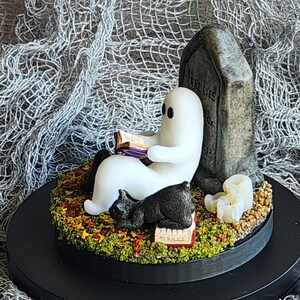 Side view to show detail. Ghost sitting beside gravestone reading a book with 2 black cats playing in the fall leaves. Halloween decor that will glow in the dark and has flickering candles.