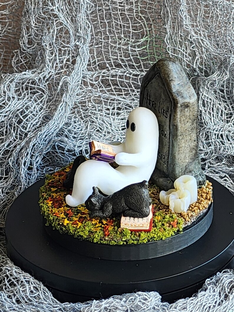 Side view to show detail. Ghost sitting beside gravestone reading a book with 2 black cats playing in the fall leaves. Halloween decor that will glow in the dark and has flickering candles.