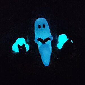 Glow in the dark photo. The ghost, candles, and cat eyes glow. Ghost sitting beside gravestone reading a book with 2 black cats playing in the fall leaves. Halloween decor that will glow in the dark and has flickering candles.