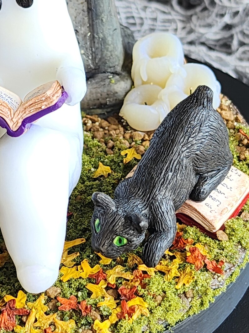Close up of manx black cat on a book ready to pounce and play in the autumn leaves. Ghost sitting beside gravestone reading a book with 2 black cats playing in the fall leaves. Halloween decor that will glow in the dark and has flickering candles.