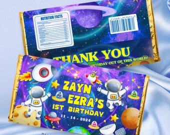 May include: A blue and purple galaxy-themed chocolate bar wrapper with a rocket, astronaut, and planets. The text on the wrapper says "Zayn Ezra's 1st Birthday 11-14-2024" and "Thank You Birthday Out of This World!"