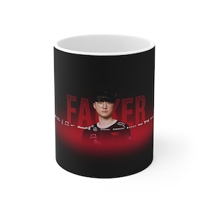 Può includere: Tazza in ceramica nera con un'immagine rossa e bianca di un uomo con gli occhiali e una camicia nera e rossa. La parola "FAKER" è scritta in grandi lettere rosse sull'immagine.
