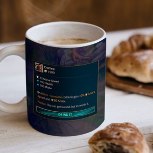 Könnte beinhalten: Weiße Keramik-Kaffeetasse mit Henkel, gefüllt mit Kaffee. Die Tasse hat eine dunkelblaue Grafik mit dem Text "Coffee" und "DRINK IT". Die Tasse steht auf einer weißen Oberfläche mit Gebäck im Hintergrund.