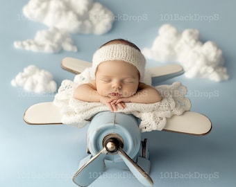 May include: A newborn baby wearing a white knitted hat sleeps on a blue and white toy aeroplane prop. The aeroplane is surrounded by white cotton clouds against a light blue background.