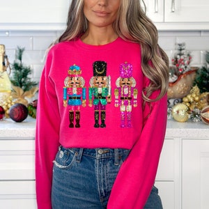 May include: A hot pink sweatshirt featuring three decorative nutcracker figures. The nutcrackers are in shades of blue, green, and pink with gold and black accents. The sweatshirt is a crew neck style.