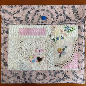 Pode incluir: Uma bolsa de tecido rosa com um padrão floral e um fecho de botão azul. A frente apresenta um design de patchwork com renda, flores bordadas e o texto "Belle Fleur". A bolsa é um acessório decorativo.