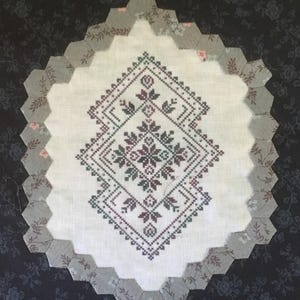 May include: A white fabric square with a geometric cross-stitch design in shades of green, brown, and black. The square is surrounded by a border of gray fabric with a floral pattern.