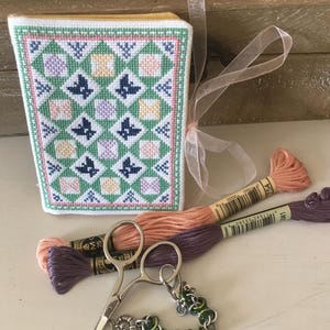 May include: A small, cross-stitched book cover with a geometric pattern in green, pink, yellow, and blue. Two skeins of embroidery floss in peach and purple, small silver scissors, and a green and silver chain bracelet are also present.