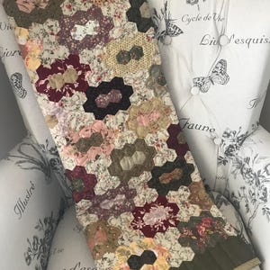 Könnte beinhalten: Eine handgefertigte Steppdecke mit einem floralen Sechseckmuster in Rosa-, Burgunder-, Creme- und Olivgrüntönen. Die Steppdecke ist über einem weißen Sessel mit Blumen- und Schmetterlingsdesign drapiert.