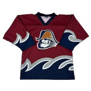 May include: Maroon hockey jersey with a V-neck and long sleeves. The jersey features a team logo of a pirate with a tricorn hat. The sleeves and bottom hem have a wave design in navy, gray, and white.