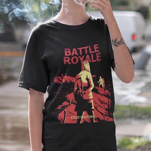 May include: Black t-shirt with a graphic print of a woman in a red jacket walking on a red and black landscape. The text "BATTLE ROYALE" is printed in yellow above the graphic and "KINJI FUKASAKU" is printed in yellow below the graphic.