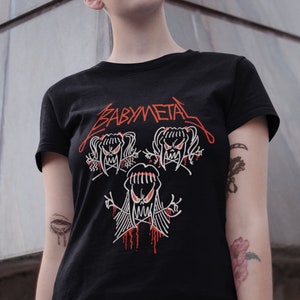Pode incluir: Uma camiseta preta com um gr&aacute;fico branco de tr&ecirc;s figuras de desenhos animados com acentos vermelhos. O texto "BABYMETAL" est&aacute; impresso em vermelho na camiseta.