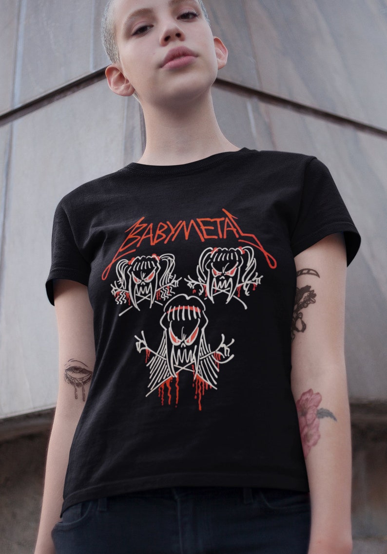 Pode incluir: Uma camiseta preta com um gr&aacute;fico branco de tr&ecirc;s figuras de desenhos animados com acentos vermelhos. O texto "BABYMETAL" est&aacute; impresso em vermelho na camiseta.