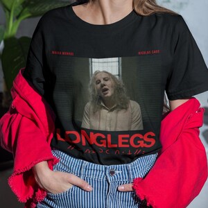 Puede incluir: Camiseta negra con un gr&aacute;fico de Nicolas Cage de la pel&iacute;cula "Longlegs". El gr&aacute;fico est&aacute; en un estilo de foto en blanco y negro con el t&iacute;tulo "Longlegs" en texto rojo.