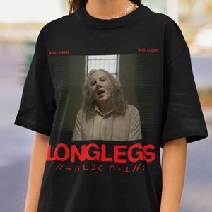 Puede incluir: Camiseta negra con un gr&aacute;fico rojo de Nicolas Cage de la pel&iacute;cula "Longlegs" con el texto "Longlegs" y "// - _ L O N G L E G S - _ //" impreso en la parte delantera.