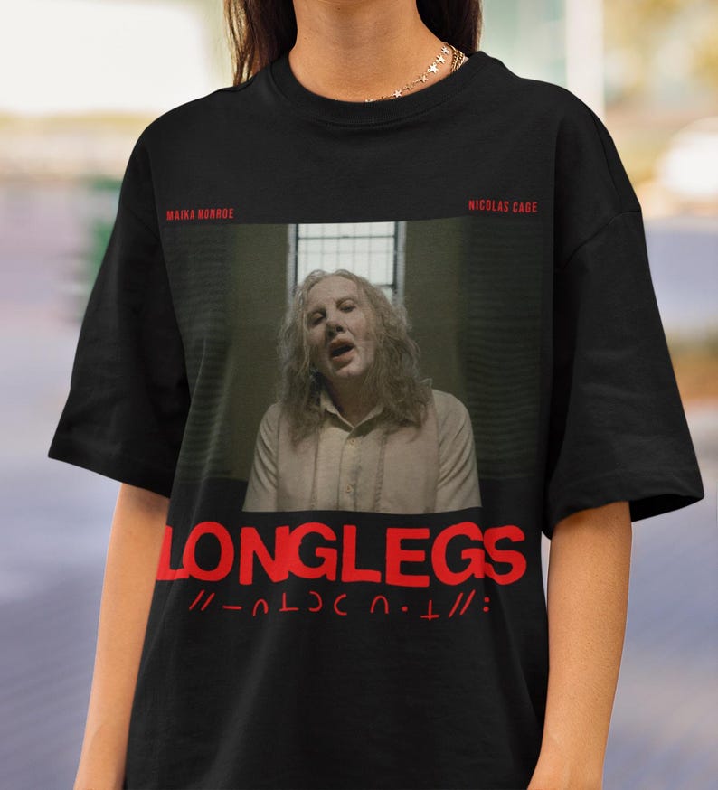 Puede incluir: Camiseta negra con un gr&aacute;fico rojo de Nicolas Cage de la pel&iacute;cula "Longlegs" con el texto "Longlegs" y "// - _ L O N G L E G S - _ //" impreso en la parte delantera.