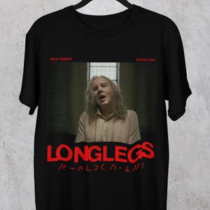 Puede incluir: Camiseta negra con un gr&aacute;fico rojo de una mujer con el pelo largo y el texto "LONGLEGS //-ALDCN.T//:"  El texto "MAIKA MONROE" y "NICOLAS CADE" aparecen en rojo m&aacute;s peque&ntilde;o encima del gr&aacute;fico.