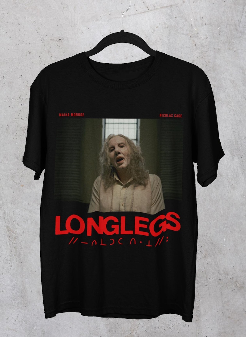 Puede incluir: Camiseta negra con un gr&aacute;fico rojo de una mujer con el pelo largo y el texto "LONGLEGS //-ALDCN.T//:"  El texto "MAIKA MONROE" y "NICOLAS CADE" aparecen en rojo m&aacute;s peque&ntilde;o encima del gr&aacute;fico.