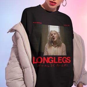 Puede incluir: Camiseta negra con un gr&aacute;fico de una mujer con el pelo largo y una camisa blanca. El texto "LONGLEGS" est&aacute; impreso en rojo en la camiseta. Los textos "MAIKA MONROE" y "NICHOLAS CAGE" est&aacute;n impresos en blanco en la camiseta.