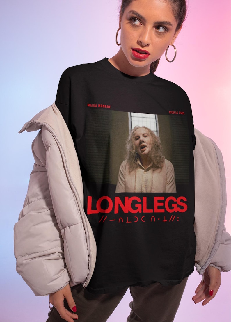 Puede incluir: Camiseta negra con un gr&aacute;fico de una mujer con el pelo largo y una camisa blanca. El texto "LONGLEGS" est&aacute; impreso en rojo en la camiseta. Los textos "MAIKA MONROE" y "NICHOLAS CAGE" est&aacute;n impresos en blanco en la camiseta.