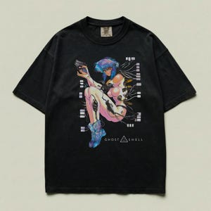 May include: A black short-sleeve t-shirt featuring a graphic print of a cyborg character with blue hair holding a handgun. The character is connected to various wires and cables. The text GHOST IN THE SHELL is printed in white below the graphic.