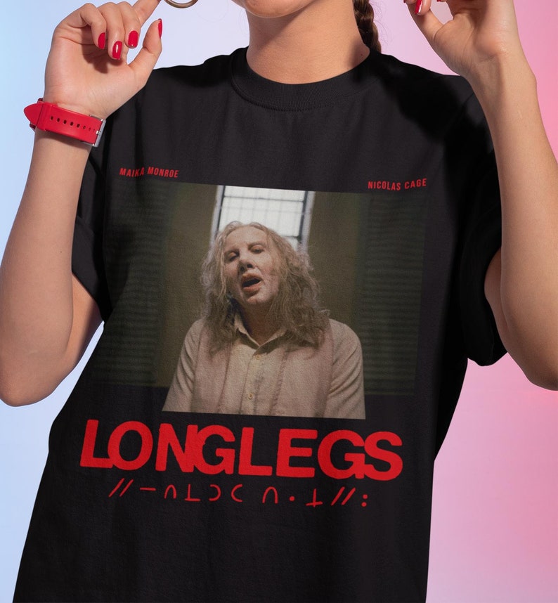 Puede incluir: Camiseta negra con un gr&aacute;fico rojo de Nicolas Cage de la pel&iacute;cula "Longlegs" con el texto "LONGLEGS //-OLC.U.T///:" en rojo.