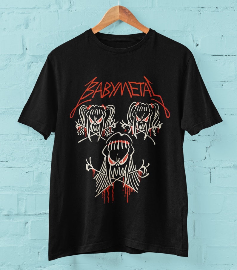 Pode incluir: Camiseta preta com um logotipo "BABYMETAL" vermelho e um gr&aacute;fico branco de tr&ecirc;s cr&acirc;nios de desenho animado com cabelo comprido.