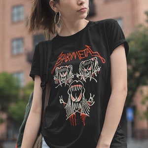 Pode incluir: Uma camiseta preta com um gr&aacute;fico branco de tr&ecirc;s cr&acirc;nios de desenhos animados com o texto "BABYMETAL" em vermelho.